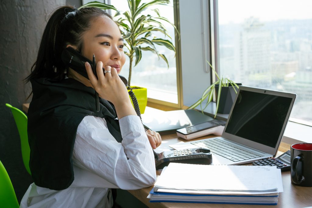Eine Frau sitzt an ihrem Schreibtisch und telefoniert mit einem Festnetz Telefon. Sie hält einen Stift in der Hand und lächelt beim Telefonat